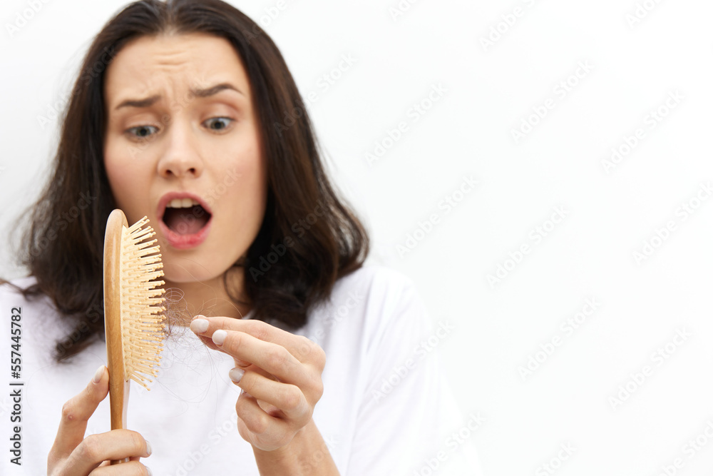 a very upset, scared, sad woman with long black hair stands in a white T-shirt on a light background and cleans a comb from fallen hair, expressing negative emotions on her face a very upset, scared, sad woman with long black hair stands in a white T-shirt on a light background and cleans a comb from fallen hair, expressing negative emotions on her face