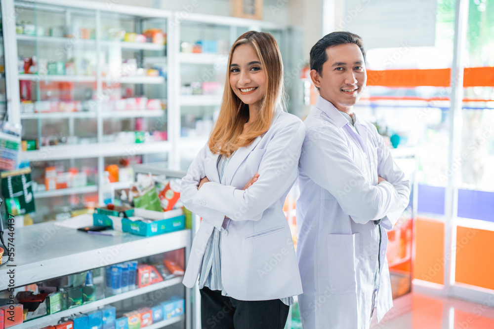 Smiling female pharmacist and male pharmacist with crossed hands standing inside drugstore