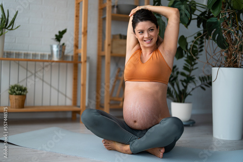 Beautiful smiling pregnant woman sitting on fitness mat and exercising during late pregnancy. Stretching with arms above the head, looking satisfied and relaxed.
