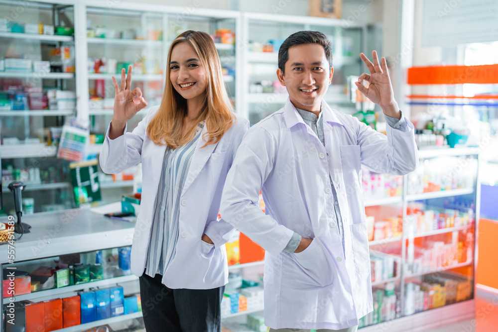 Two asian female and male pharmacists smiling with okay hand gesture at the drugstore