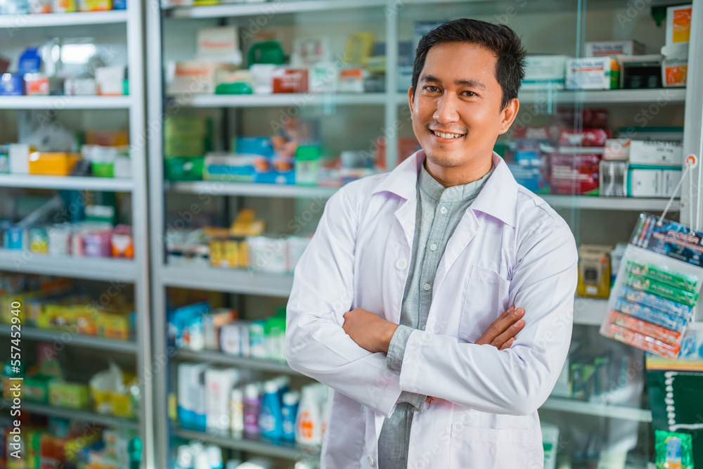 male pharmacist in uniform smiling with crossed hands on pharmacy display case background