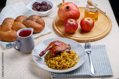 Rosh Hashanah next to pomegranate honey apples wine and challah on a white table and candles next to a duck's leg and spelt in a plate.