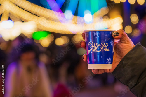 A hand holding a winter wonderland cup with blurred background of the Hyde Park Winter wonderland festival in London © Peppygraphics