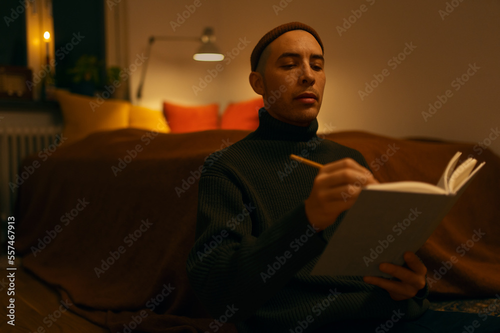 A caucasian man wearing a knitted sweater and a beanie sitting in a bedroom in a warm light writing in a notebook. A caucasian man wearing a knitted sweater and a beanie sitting in a bedroom in a warm light writing in a notebook.