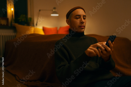 A caucasian man wearing a knitted sweater and a beanie sitting in a bedroom in a warm light holding a phone.