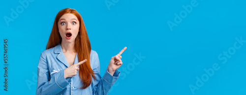 Surprised and shocked redhead female teenager living dormitory with roommate gossiping about neigbours, drop jaw gasping speechless staring camera, pointing left, blue background photo