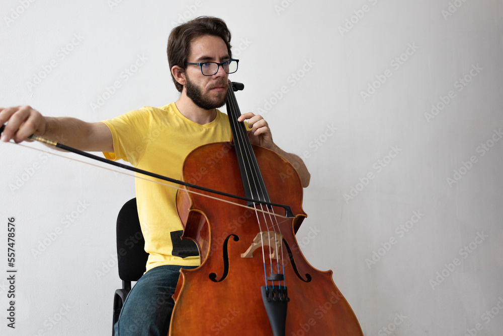 young cello player practincing in a white room, young man playing violoncello at home practicing music teaching from home young cello player practincing in a white room, young man playing violoncello at home practicing music teaching from home