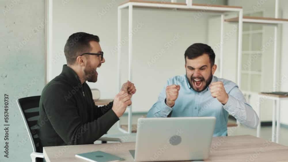 Two young men are happy and hugging while sitting at the laptop in the office Two young men are happy and hugging while sitting at the laptop in the office