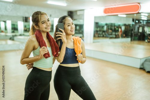 two beautiful asian women carrying towels during break and selfie using cellphones in fitness center room