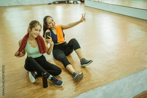 Two women sitting relaxing taking selfies together using cellphones after a workout at the gym
