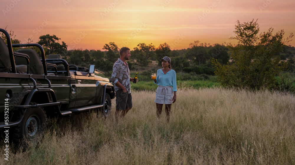 Asian women and European men on safari game drive in South Africa Kruger national park. a couple of men and women on safari. Tourists in a jeep looking sunset with drinks on safari - obrazy, fototapety, plakaty 