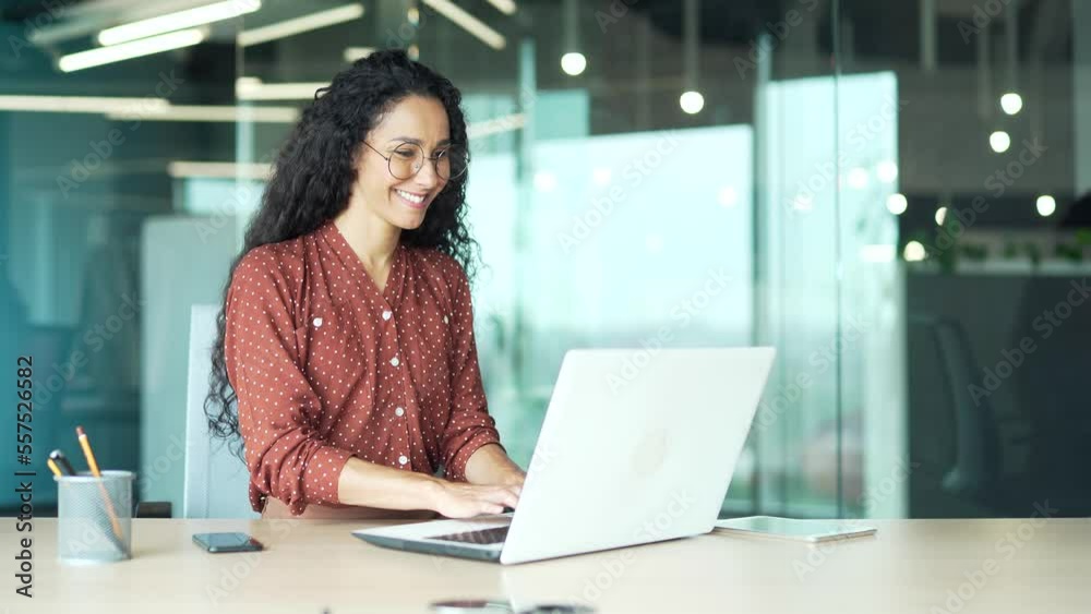 Curly brunette employee works in a modern office at workplace at a laptop computer focused and confident woman programmer web developer writing code typing programming business female worker in casual