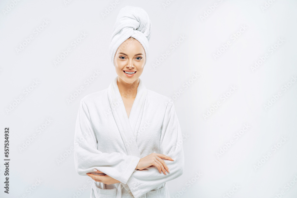 Beautiful smiling young woman has soft healthy skin after taking shower, wears bath robe and towel wrapped on head, enjoys spare time at home, isolated over white background. Wellness concept. - obrazy, fototapety, plakaty 