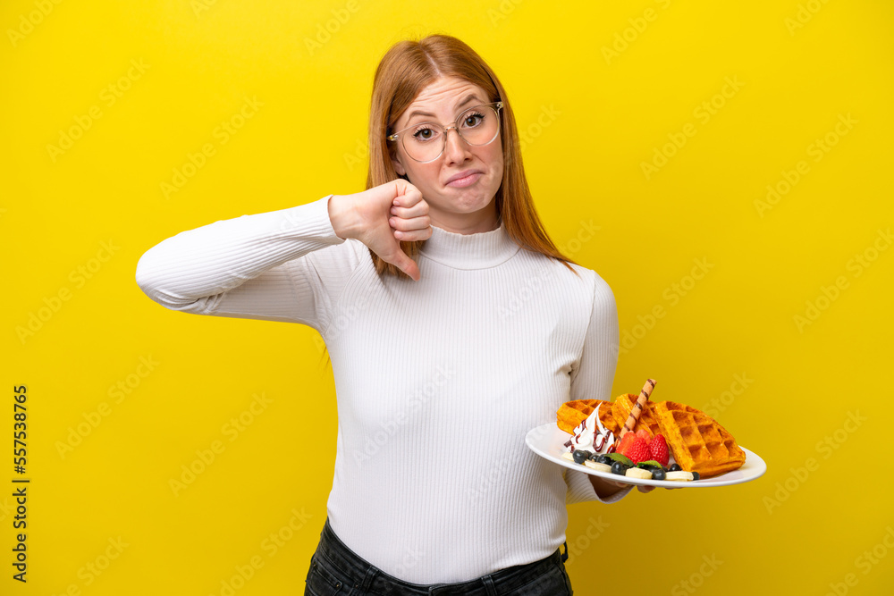Young redhead woman holding waffles isolated on yellow background showing thumb down with negative expression Young redhead woman holding waffles isolated on yellow background showing thumb down with negative expression