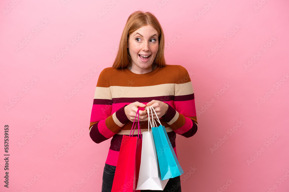 Young redhead woman isolated on pink background holding shopping bags and surprised Young redhead woman isolated on pink background holding shopping bags and surprised