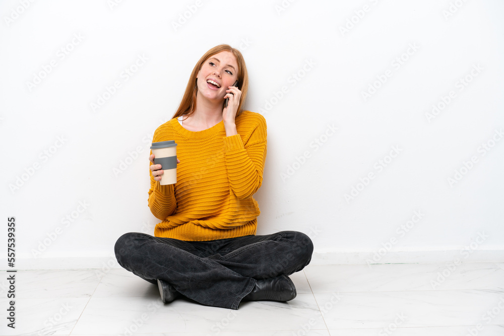 Young redhead woman sitting on the floor isolated on white background holding coffee to take away and a mobile Young redhead woman sitting on the floor isolated on white background holding coffee to take away and a mobile
