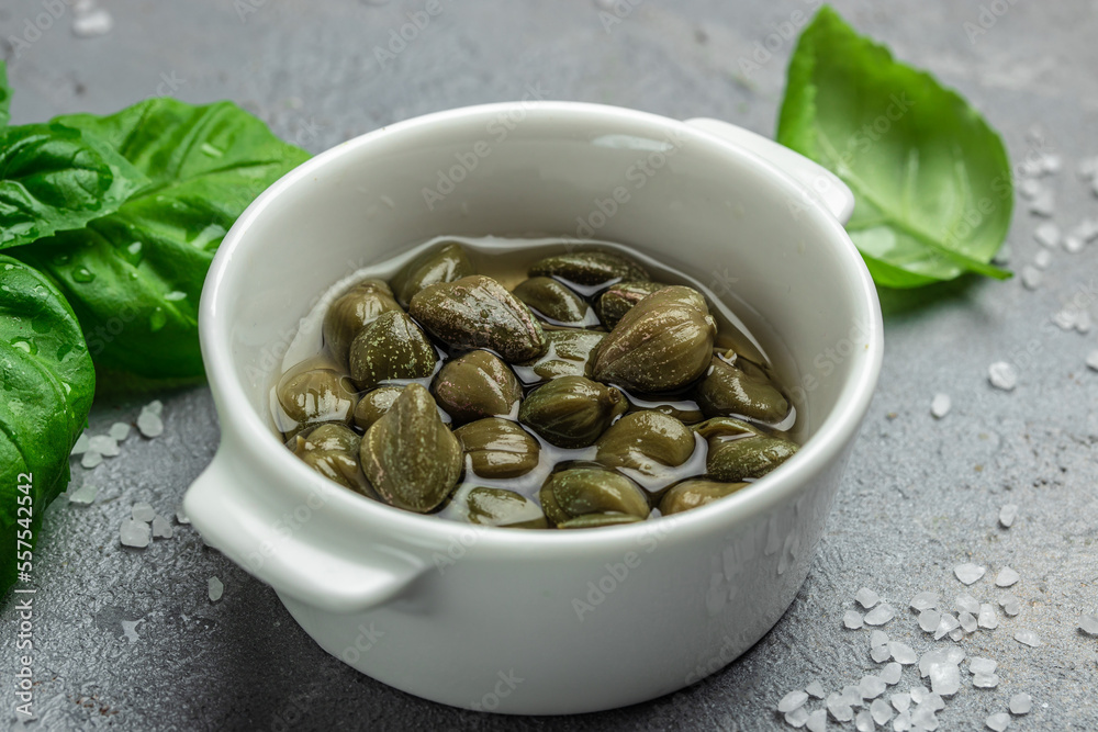 Marinated capers with fresh basil leaves in a small bowl, on light wooden background. Food recipe background. Close up Marinated capers with fresh basil leaves in a small bowl, on light wooden background. Food recipe background. Close up