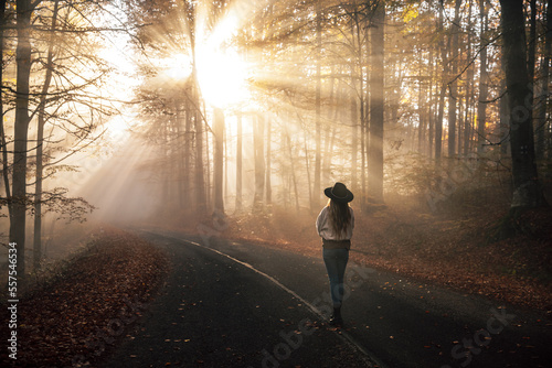 Young woman standing in the rays of the morning sun in a leafy autumn forest and enjoying the magical view. 