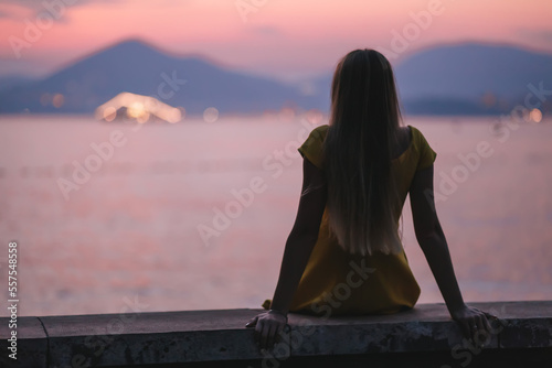Fotobehang A young beautiful woman in a dress sits on the beach at sunset and looks at the setting sun, Lonely girl dreams of love