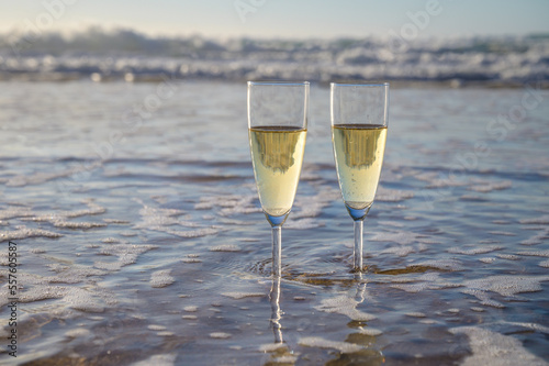 Glasses of cava or champagne sparkling wine on white sandy ocean beach with water waves on sunset in sunlights