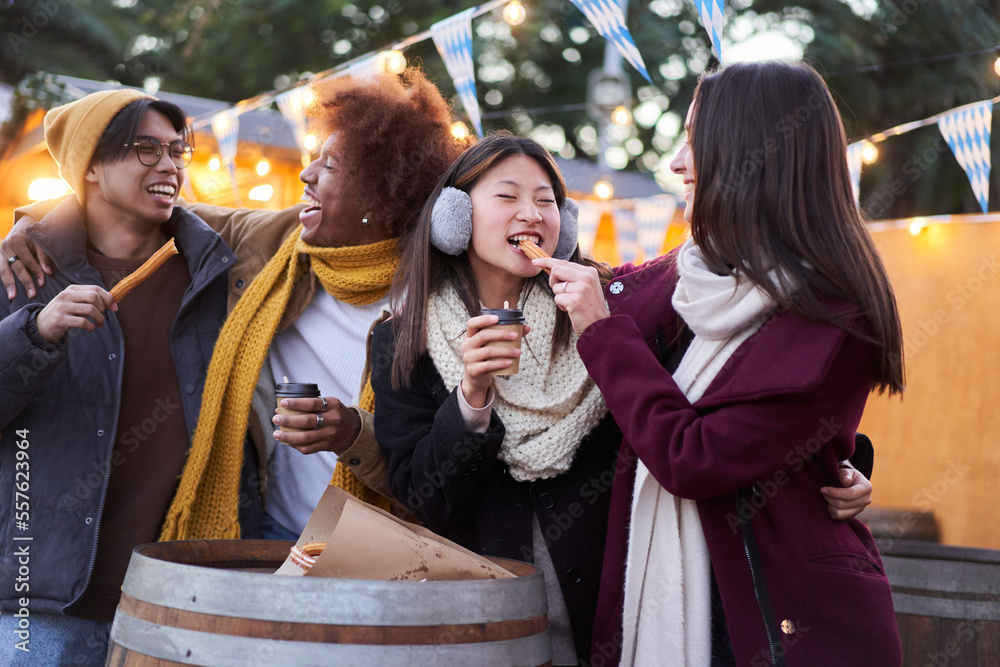 Happy friends eating chocolate with churros street food at the city in the night outdoors. Tourist group of four people having fun in winter time. One girl feeds the other. Friendship concept. Happy friends eating chocolate with churros street food at the city in the night outdoors. Tourist group of four people having fun in winter time. One girl feeds the other. Friendship concept.
