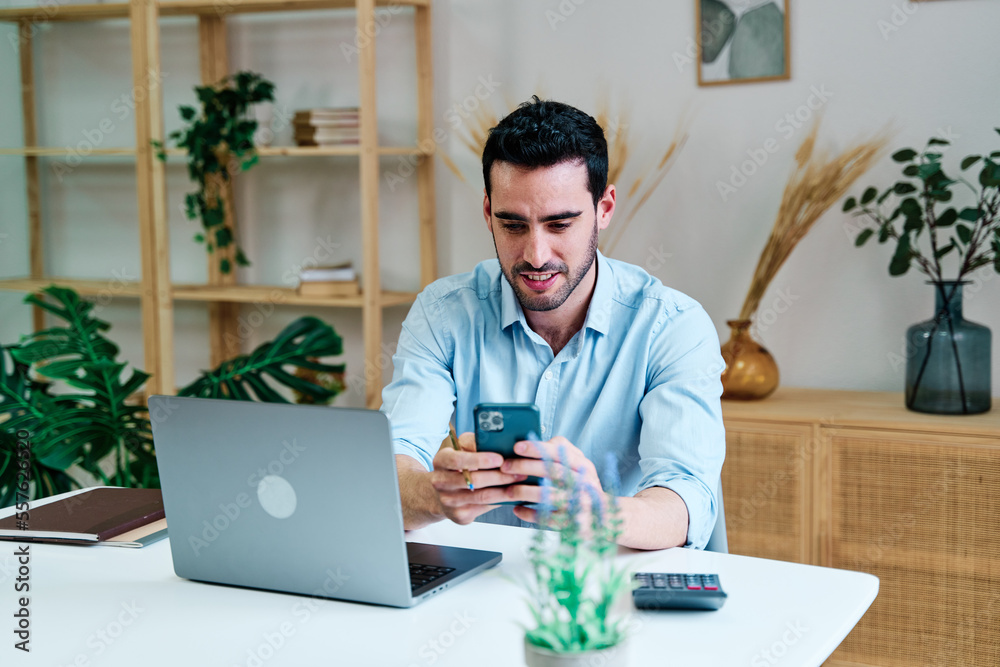 Young male freelancer in casual clothes sitting at table with laptop and browsing mobile phone while working on remote project at home Young male freelancer in casual clothes sitting at table with laptop and browsing mobile phone while working on remote project at home