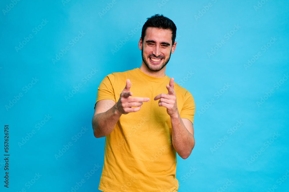 Cheerful young male in yellow shirt smiling and showing thumb up gesture while standing against blue background