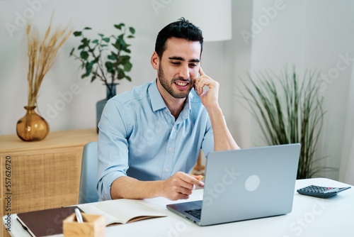 Positive male freelancer in casual clothes sitting at table with laptop and smartphone while working on remote project at home