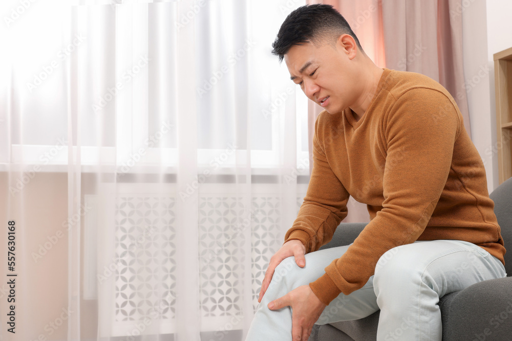 Asian man suffering from knee pain on sofa indoors Asian man suffering from knee pain on sofa indoors