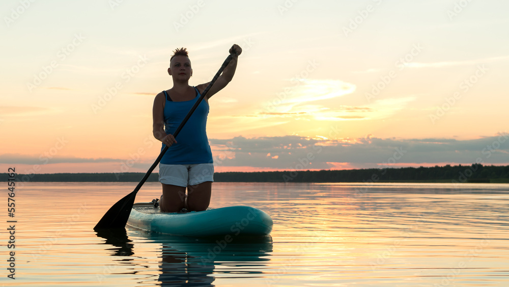 A woman on a sapboard on her knees with an oar at sunset against the sky floats in the water of the lake.
