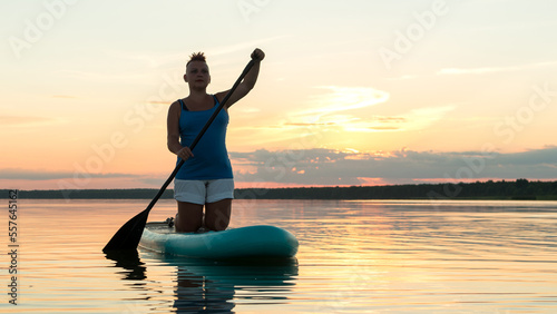 A woman on a sapboard on her knees with an oar at sunset against the sky floats in the water of the lake. © finist_4
