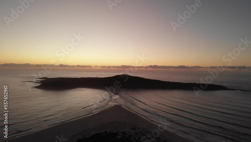 Drone video flying toward Shark Island, Final Bay, as the sun rises behind it. Port Stephens lighthouse silhouetted against the bright sky.