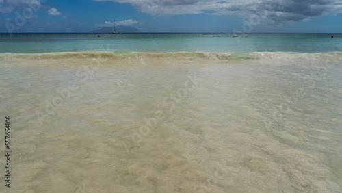 A wave rolls onto the shore. The water mixes with the sand of the beach. Yachts in the turquoise ocean. The silhouette of the island on the horizon. Clouds in the blue sky. Seychelles. Mahe. 