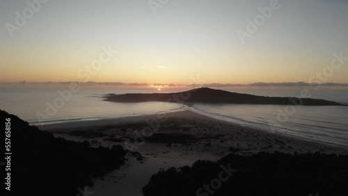 Drone video flying toward Shark Island, Final Bay, as the sun rises behind it. Port Stephens lighthouse silhouetted against the bright sky.