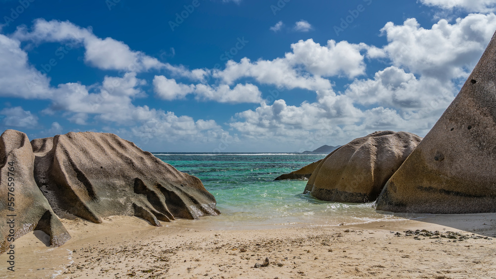 Naklejka premium Picturesque granite rocks with rounded folded slopes on the beach of a tropical island. The waves of the turquoise ocean spread over the sand. Clouds in the blue sky. Seychelles. La Digue. 