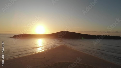 Drone video flying toward Shark Island, Final Bay, as the sun rises behind it. Port Stephens lighthouse silhouetted against the bright sky.