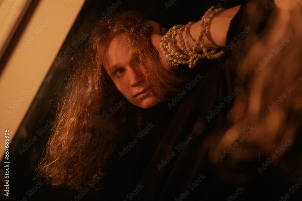 Portrait of young man with long blond curly hair under the light of lamp and candles in the mirror. Solitude, bright loneliness, creativity, thoughtfulness Portrait of young man with long blond curly hair under the light of lamp and candles in the mirror. Solitude, bright loneliness, creativity, thoughtfulness