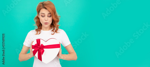 surprised woman hold heart present box on blue background. Woman isolated face portrait, banner with mock up copyspace. © Olena
