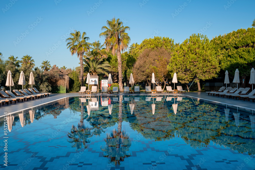 Empty deck chairs and folded parasols surrounded with trees and clear blue sky in the background reflecting on swimming pool at tourist resort