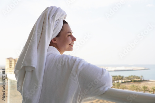 Woman in bathrobe and towel stands on hotel balcony and looks into the distance