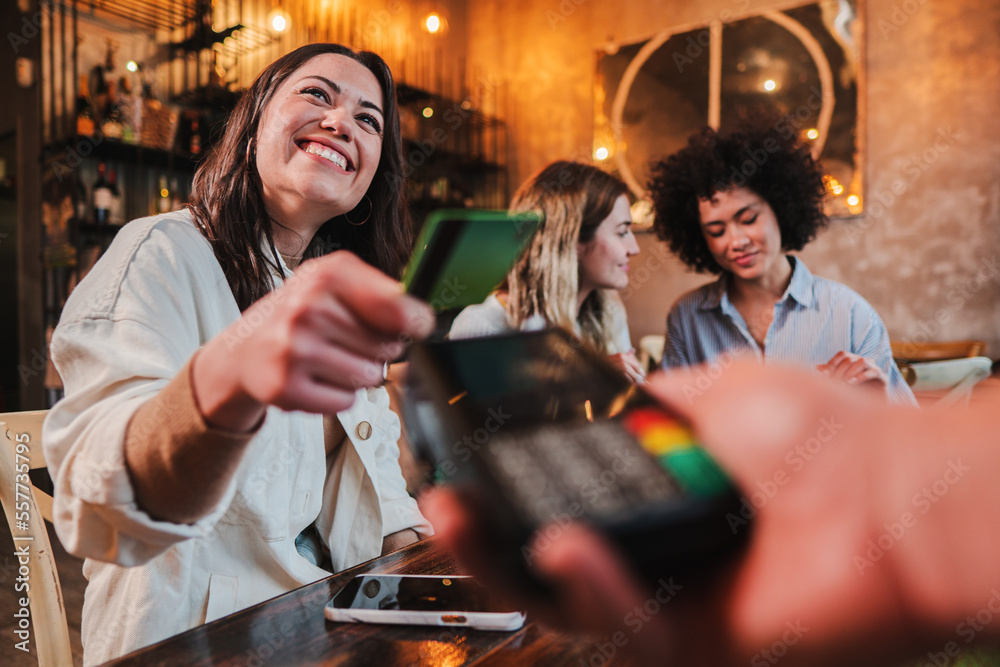 Happy young woman paying bill with a contactless credit card in a restaurant. Female smiling holding a creditcard and giving a payment transaction to the cashier. High quality photo - obrazy, fototapety, plakaty 