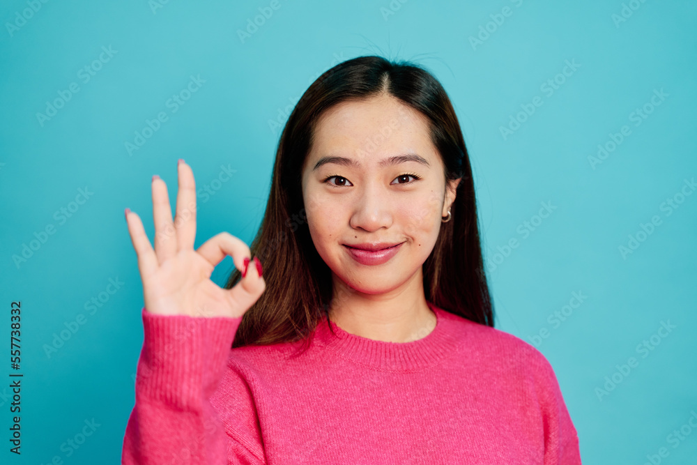 Positive young female in pink sweater smiling and showing OK gesture while looking at camera on blue background in studio Positive young female in pink sweater smiling and showing OK gesture while looking at camera on blue background in studio