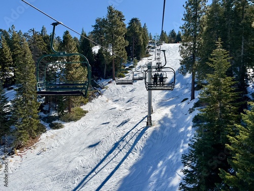 Rear view of unidentified skiers and snowboarders on chairlift going up snow covered mountain, past pine trees, at Big Bear, California photo