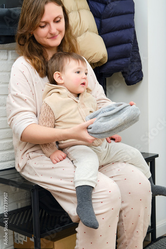 Mother puts a warm scarf on the toddler baby sitting in the home hallway. Woman mom dressing warm clothes on child for winter walk in cold weather. Kid aged one year and three months