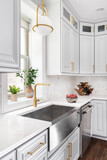 A kitchen sink detail shot with a gold pendant light, stainless steel apron sink, white cabinets, and herringbone marble backsplash.