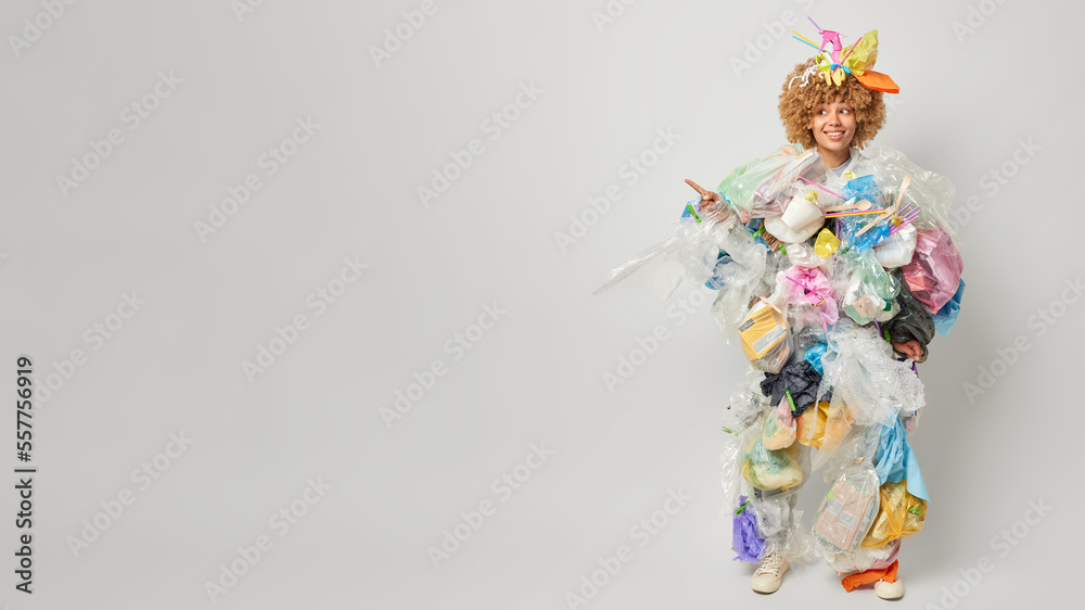 Optimistic female volunteer covered with plastic garbage takes care of environment points index finger aside demonstrates place for your advertisement isolated over grey background. Recycling concept Optimistic female volunteer covered with plastic garbage takes care of environment points index finger aside demonstrates place for your advertisement isolated over grey background. Recycling concept