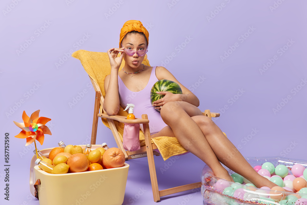 Lovely young lady with slender legs wears swimsuit sunglasses poses with watermelon on deck chair spends summer vacations at seaside uses portable fridge and inflated pool isolated over purple wall