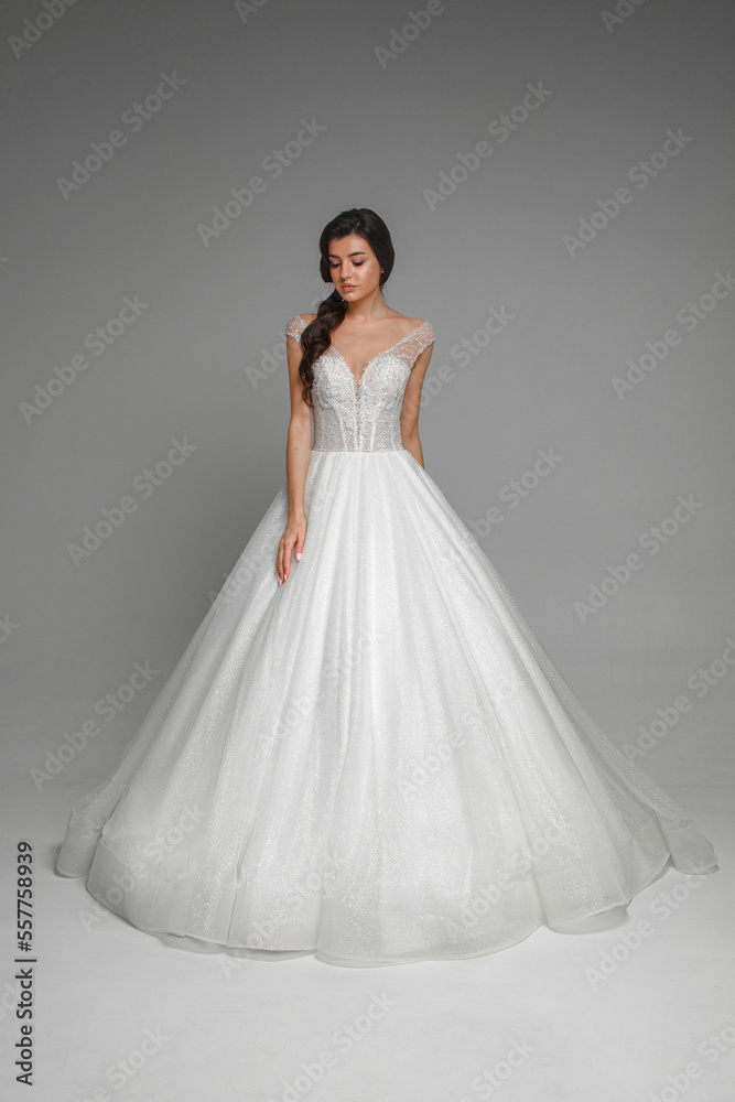 Pretty young caucasian brunette bride with make-up and pony tail hairstyle in long wedding ball gown dress standing and posing in grey interior.
