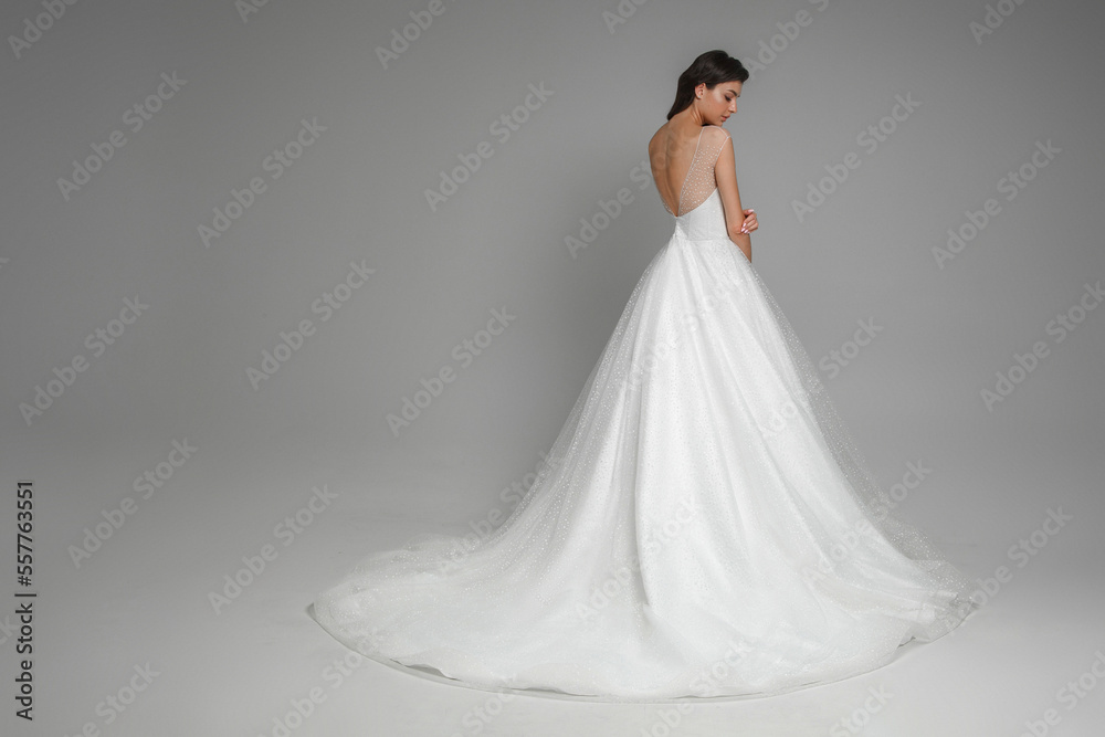 Portrait from the back of beautiful caucasian brunette bride in the beautiful white wedding dress standing in the grey studio. Gorgeous beautiful female on a isolated background.