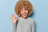 Friendly cheerful European woman with curly hair waves hello and smiles broadly wears casual grey jumper greets someone being in good mood isolated over blue background. Hey nice to meet you